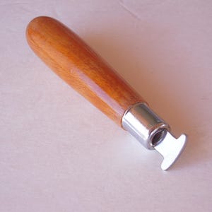 May include: A wooden handled tool with a polished finish. The handle is a light brown color. The tool has a silver-colored metal end with a curved, flat blade. The tool is likely used for leatherworking or similar crafts.