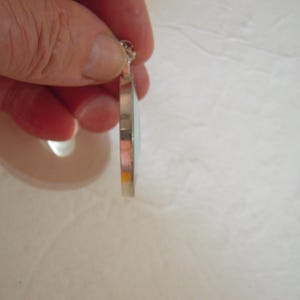 May include: A silver pendant with a light blue and orange design. The pendant is held by a hand, showcasing its side profile. The pendant has a circular shape and a small loop at the top for a chain.