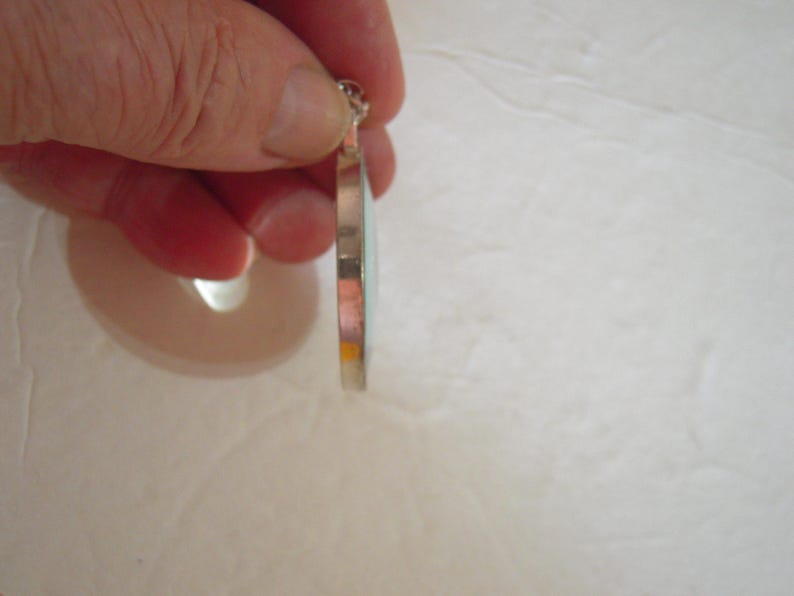 May include: A silver pendant with a light blue and orange design. The pendant is held by a hand, showcasing its side profile. The pendant has a circular shape and a small loop at the top for a chain.