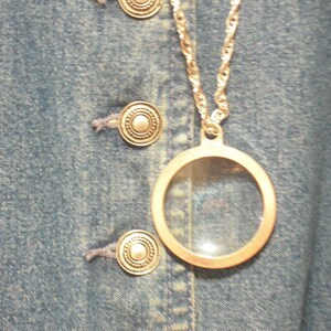 May include: A silver-toned necklace with a circular pendant, featuring a clear glass center. The necklace hangs against a blue denim background with silver buttons.