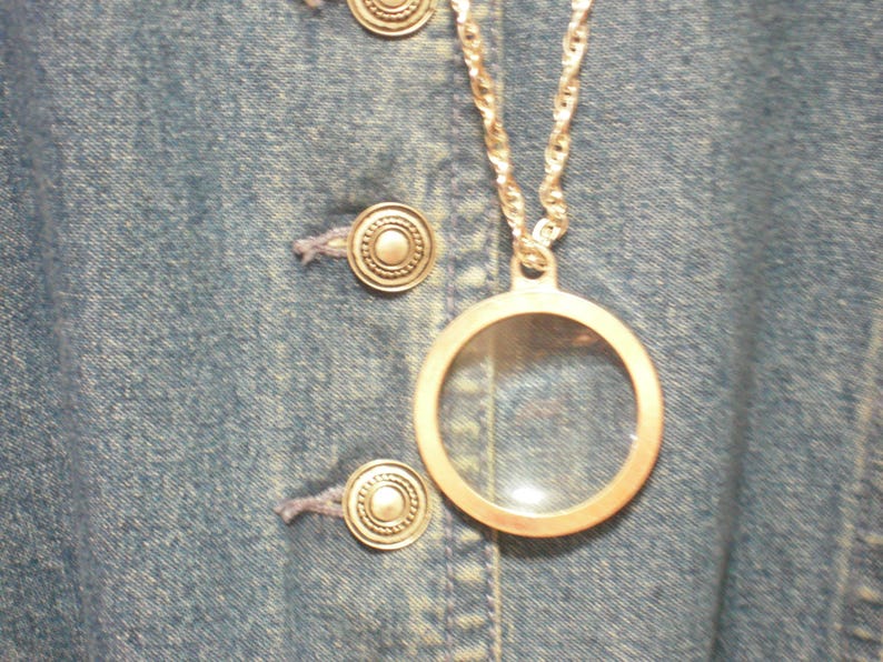 May include: A silver-toned necklace with a circular pendant, featuring a clear glass center. The necklace hangs against a blue denim background with silver buttons.