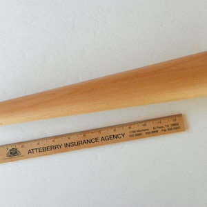 May include: A wooden tapered furniture leg, approximately 12 inches long, with a diameter of 1.5 inches at the widest point. The leg is made of light-colored wood and is shown next to a ruler for scale.