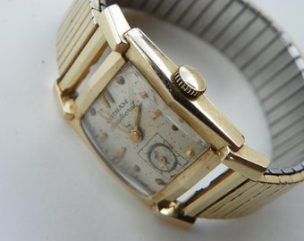 May include: A gold-toned wristwatch with a square face and a metal band. The watch face has a white background with black Roman numeral hour markers and black hands. The watch face also has the text "Gotham  waterproof" printed on it.