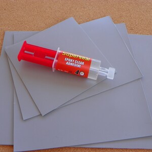 May include: Four grey square sheets of material with a syringe of Superseal Epoxy Clear Adhesive on top. The syringe is red and white with the words "Superseal Epoxy Clear Adhesive" printed on the side.