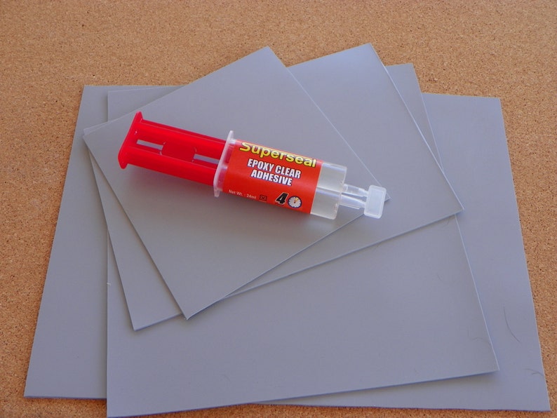 May include: Four grey square sheets of material with a syringe of Superseal Epoxy Clear Adhesive on top. The syringe is red and white with the words "Superseal Epoxy Clear Adhesive" printed on the side.