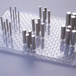 May include: A clear plastic pegboard with rows of silver metal pegs of varying lengths. The pegs are arranged in a random pattern.