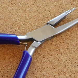 May include: A pair of stainless steel jewelry pliers with blue handles. The pliers have a rounded nose and are designed for bending and shaping wire. The tool is shown against a corkboard background.
