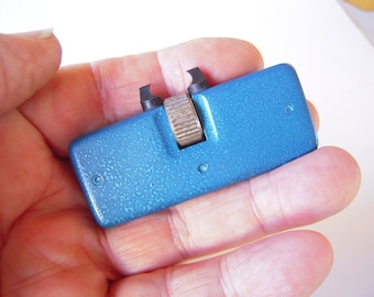 Puede incluir: Un abridor de caja de reloj de metal azul con una perilla plateada y dos puntas. El abridor se sostiene en una mano.
