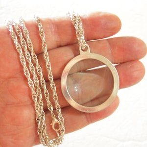 May include: Silver-toned necklace featuring a round, clear locket pendant. The pendant is suspended from a delicate chain. The locket has a simple, elegant design, perfect for holding small keepsakes. The chain is approximately 20 inches long.