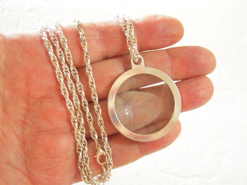 May include: Silver-toned necklace featuring a round, clear locket pendant. The pendant is suspended from a delicate chain. The locket has a simple, elegant design, perfect for holding small keepsakes. The chain is approximately 20 inches long.