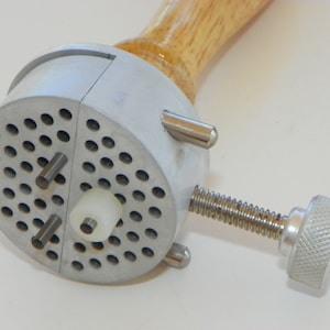 May include: A metal and wood tool with a perforated circular head. The head has several small cylindrical pins and a white plastic cylinder. A threaded screw with a textured knob extends from the side. The handle is made of light-colored wood.