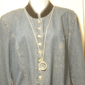 May include: A light blue denim jacket with a black velvet collar and silver buttons. A long silver necklace with a circular pendant hangs from the collar. The jacket has long sleeves and a fitted silhouette.