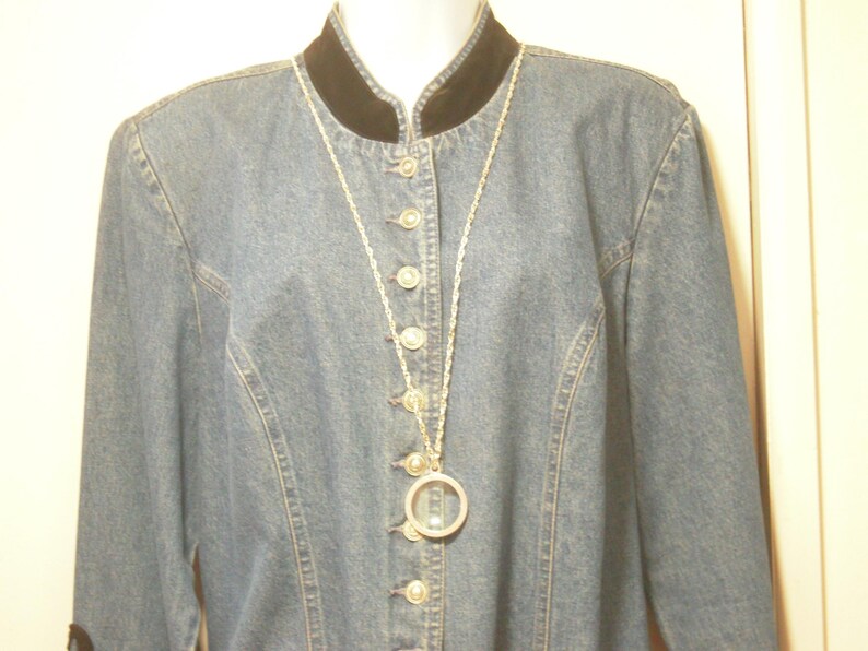 May include: A light blue denim jacket with a black velvet collar and silver buttons. A long silver necklace with a circular pendant hangs from the collar. The jacket has long sleeves and a fitted silhouette.