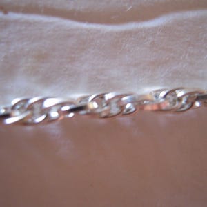 May include: A close-up of a silver chain with a twisted rope design. The chain links are interlocked, reflecting light and creating a shiny appearance. The chain is likely made of a metal such as sterling silver or stainless steel.