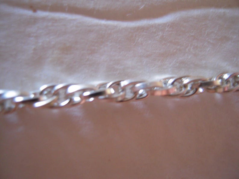 May include: A close-up of a silver chain with a twisted rope design. The chain links are interlocked, reflecting light and creating a shiny appearance. The chain is likely made of a metal such as sterling silver or stainless steel.