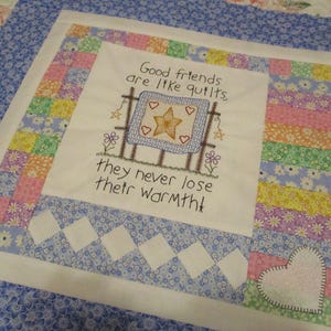 May include: A square fabric quilt block featuring a central white panel with embroidered text that reads, Good friends are like quilts, they never lose their warmth, surrounded by a colorful patchwork border of floral and pastel fabric squares.