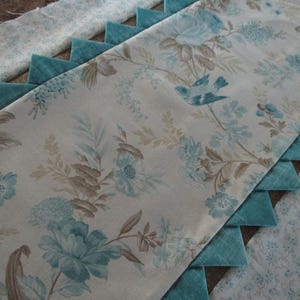 May include: A rectangular table runner featuring a cream-colored fabric with a floral and bird pattern in shades of blue and brown. The edges are decorated with a series of triangular teal fabric points, and a separate matching floral fabric strip.