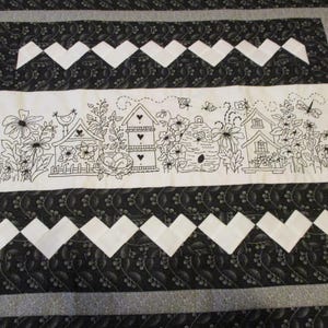 May include: A rectangular quilt featuring a central white panel with black line art of birdhouses, a beehive, flowers, and insects. This panel is bordered by black fabric with a white leaf pattern and two rows of white chevron-style heart shapes.