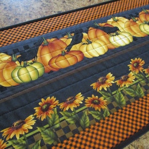 May include: A rectangular fabric table runner featuring a quilted design with a pattern of orange and yellow pumpkins, black crows, and sunflowers. The border consists of a black and orange checkered print on a dark gray background.