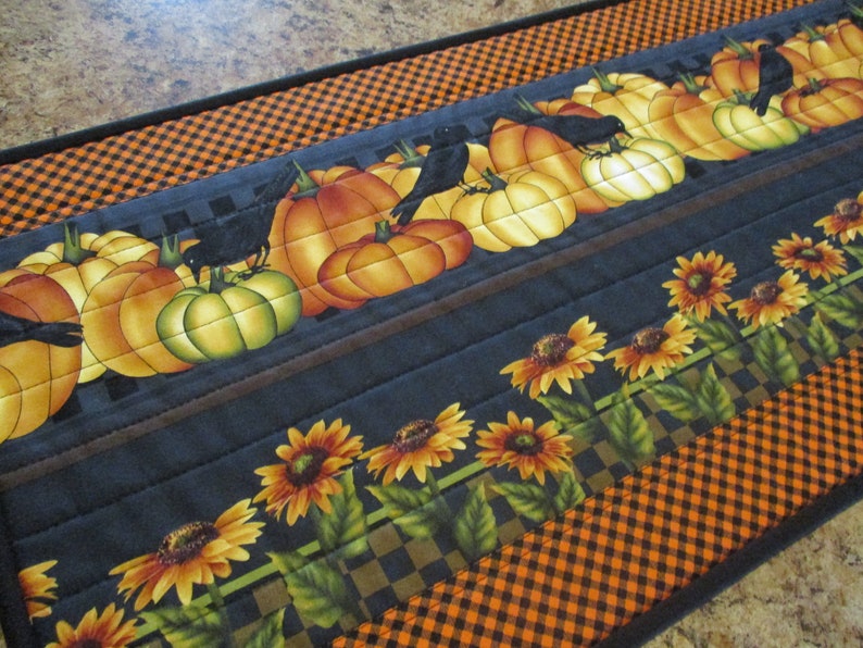 May include: A rectangular fabric table runner featuring a quilted design with a pattern of orange and yellow pumpkins, black crows, and sunflowers. The border consists of a black and orange checkered print on a dark gray background.
