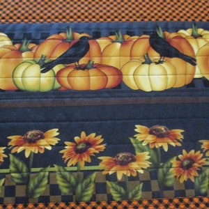 May include: A quilted fabric runner featuring a top border of orange and black gingham. The middle section displays orange pumpkins and black crows on a dark background. The bottom section shows yellow sunflowers with green leaves on a dark base.