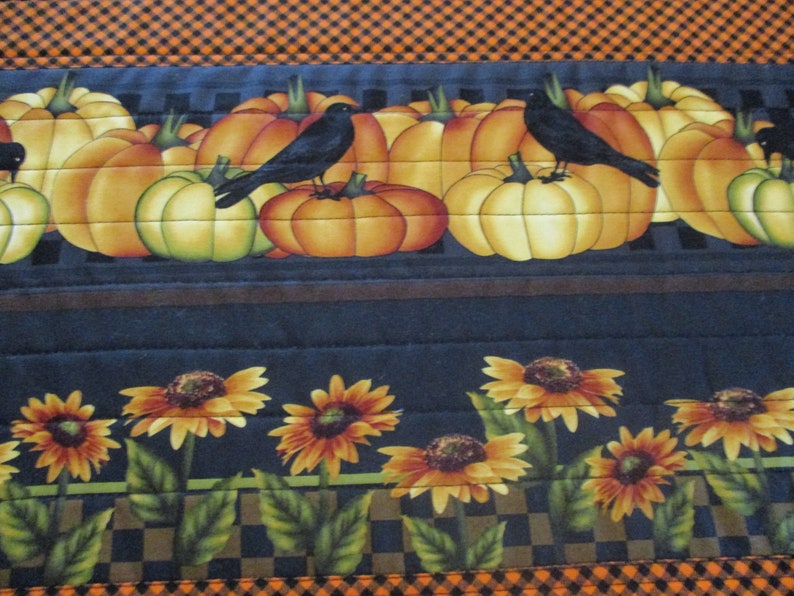 May include: A quilted fabric runner featuring a top border of orange and black gingham. The middle section displays orange pumpkins and black crows on a dark background. The bottom section shows yellow sunflowers with green leaves on a dark base.