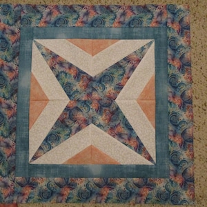 May include: A square quilt block featuring a central star pattern composed of white, peach, and blue-toned patterned fabric. The design is framed by a solid blue border and an outer border of fabric with a swirling blue and pink floral motif.