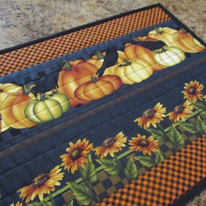 May include: A rectangular quilted table runner featuring a pattern of orange pumpkins, black crows, and yellow sunflowers. The fabric design includes a black and orange checkered border, with horizontal stitching lines across the entire surface.