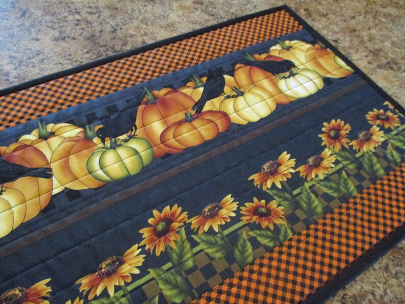 May include: A rectangular quilted table runner featuring a pattern of orange pumpkins, black crows, and yellow sunflowers. The fabric design includes a black and orange checkered border, with horizontal stitching lines across the entire surface.