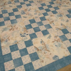 May include: A patchwork quilt featuring a repeating pattern of solid blue textured squares and cream-colored squares decorated with blue floral motifs, butterflies, and small birds. The quilt is finished with a wide blue border around the edges.