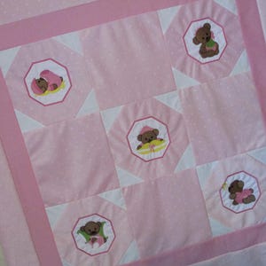 May include: A pink baby quilt with a repeating pattern of embroidered teddy bears in octagonal frames. The quilt features a light pink background with white polka dots, and a darker pink border. The teddy bears are wearing hats and dresses.