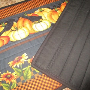 May include: A quilted fabric table runner featuring a pattern of orange pumpkins and black crows on a dark background, accented by yellow sunflowers and a black and orange checkered border. The reverse side is solid black with vertical stitching.