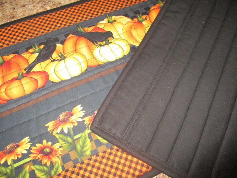 May include: A quilted fabric table runner featuring a pattern of orange pumpkins and black crows on a dark background, accented by yellow sunflowers and a black and orange checkered border. The reverse side is solid black with vertical stitching.