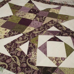 May include: Handmade quilt featuring a starburst design. The quilt showcases a patchwork of fabrics in shades of white, purple, green, and beige. The fabrics include paisley and floral patterns, creating a cozy and decorative textile.