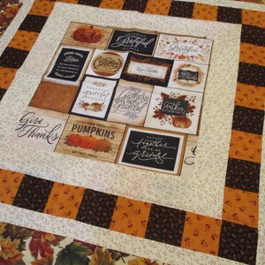 May include: A fabric quilt featuring a central panel of square blocks with autumn-themed text and pumpkin illustrations, surrounded by a border of alternating orange and dark brown squares, finished with an outer border of colorful fall leaves.