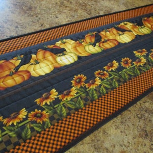 May include: A rectangular quilted table runner featuring a pattern of orange pumpkins and black crows on a dark gray background, bordered by a row of yellow sunflowers with green leaves and an outer edge of black and orange checkered fabric.