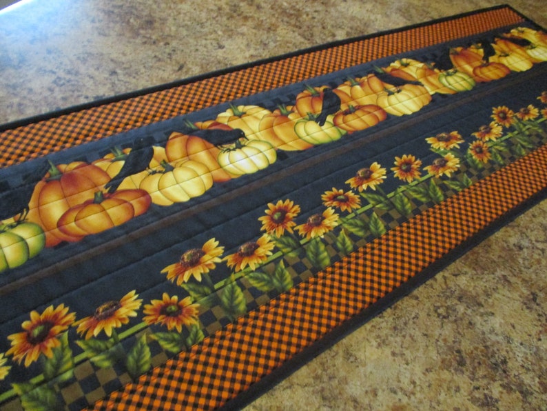 May include: A rectangular quilted table runner featuring a pattern of orange pumpkins and black crows on a dark gray background, bordered by a row of yellow sunflowers with green leaves and an outer edge of black and orange checkered fabric.