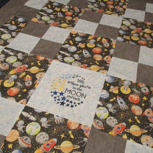 May include: A patchwork quilt featuring a space theme. The quilt has a checkerboard pattern with squares of gray, light blue, and fabric printed with planets, rockets, and astronauts. One square has the text "We love you to the MOON and back."