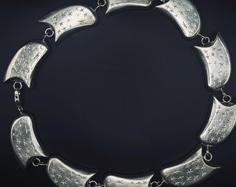 May include: A silver-toned necklace composed of ten curved, crescent-shaped links connected by small circular jump rings. Each link features a textured surface with a repeating pattern of small, etched star-like symbols across the front.