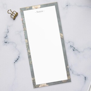 May include: A top-down view of a workspace featuring a white keyboard, a cup of coffee, and a notepad with the word "Notes-". The notepad has a floral border. Also visible are gold binder clips, colored pencils, and a small plant.