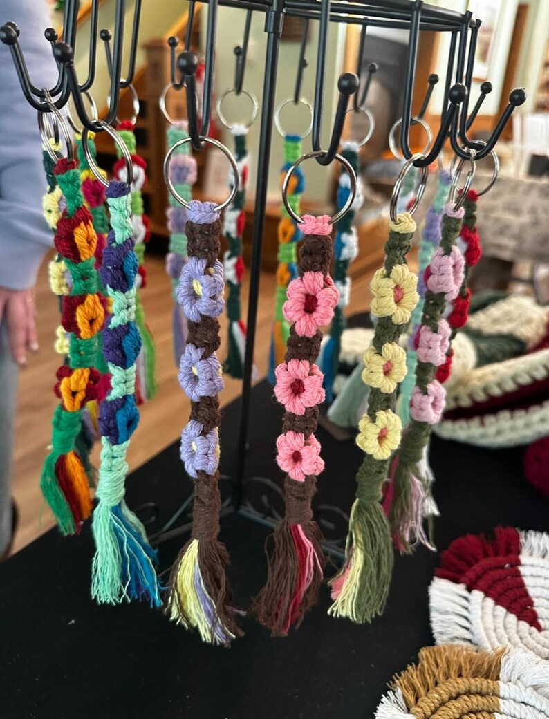 May include: Handmade macrame keychains with colorful floral designs. Each keychain features a metal ring and a tassel. The keychains are displayed on a black metal rack. The colors include pink, yellow, blue, green, and brown.