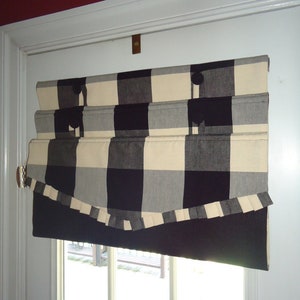 Puede incluir: Una cortina romana a cuadros negros, blancos y crema con un panel inferior negro en contraste. La cortina está montada en un marco de puerta blanco y presenta botones decorativos y un ribete con volantes. El tratamiento de la ventana está diseñado para subir y bajar.