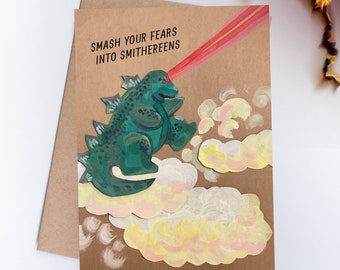 May include: A handmade greeting card with a green monster illustration shooting a red beam. The text "SMASH YOUR FEARS INTO SMITHEREENS" is printed above the monster. The card is on a brown envelope.
