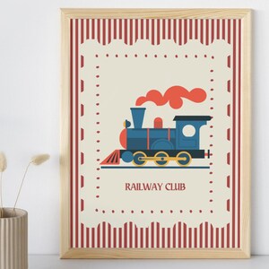May include: A framed print featuring a blue train with orange smoke, set against a cream background. The print has a red and white striped border and the words "RAILWAY CLUB" at the bottom. The frame is made of light-colored wood.
