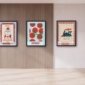 May include: Three framed art prints with black frames are displayed on a wall. The prints feature colorful illustrations: a rocket with text, tomatoes with the word "POMODORO", and a blue train with text. The wall has vertical wood paneling.
