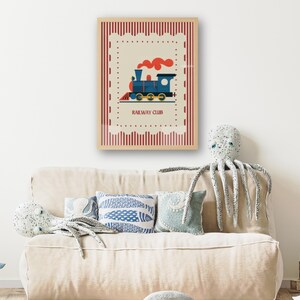 May include: A framed print featuring a blue and red cartoon train with the text "RAILWAY CLUB". The print is set against a cream background with a red border. The print is displayed above a beige sofa with decorative pillows and a stuffed octopus.