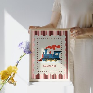 May include: A framed print featuring a blue train with red smoke, set against a cream background with a red striped border. The text "RAILWAY CLUB" is below the train illustration. The frame is light brown.