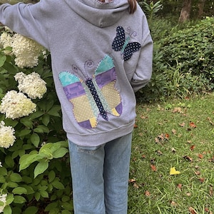 May include: A gray hoodie with two butterfly appliqués on the back. The larger butterfly is made of patchwork fabric in shades of blue, purple, and yellow. The smaller butterfly is navy blue with white polka dots. The person is wearing blue jeans.