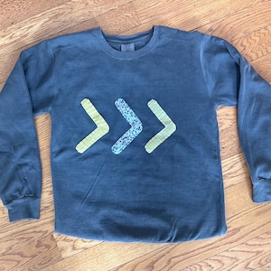 May include: A long-sleeved, crew neck shirt in a muted blue-gray color. Three chevron-shaped appliqués are centered on the chest. The outer chevrons are a light yellow, and the center chevron has a blue floral pattern.