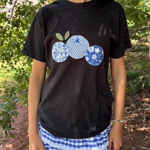 May include: Black t-shirt with a blueberry design. The design features three circular blueberries in blue and white patterned fabrics, with green leaf accents. The model is also wearing a blue and white gingham skirt.